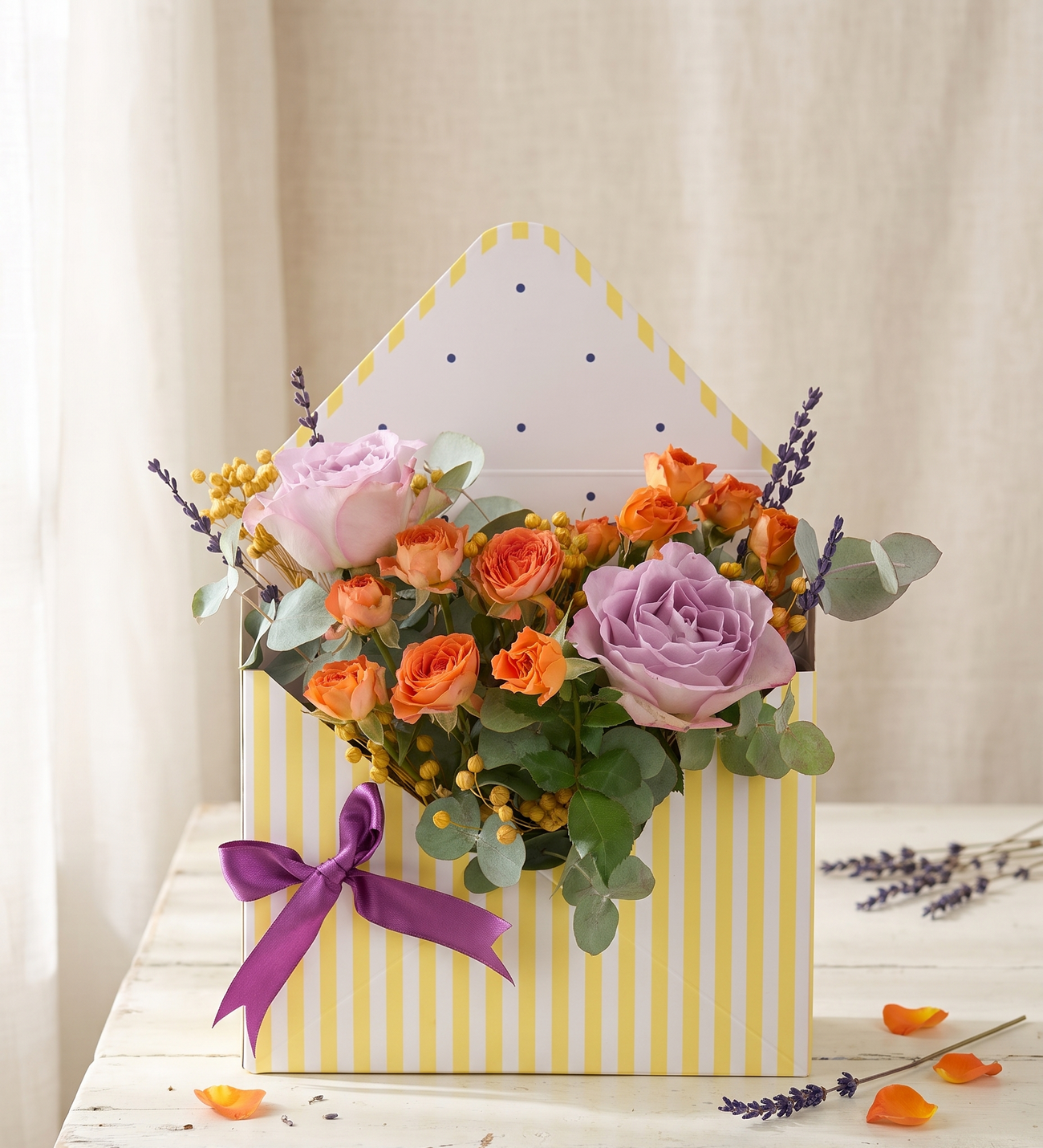 Lilac & Orange Roses in a Striped Envelope Box, Lilac garden roses and orange spray roses with yellow craspedia, lavender sprigs and eucalyptus, presented in a yellow-striped envelope-style box with a purple ribbon.
