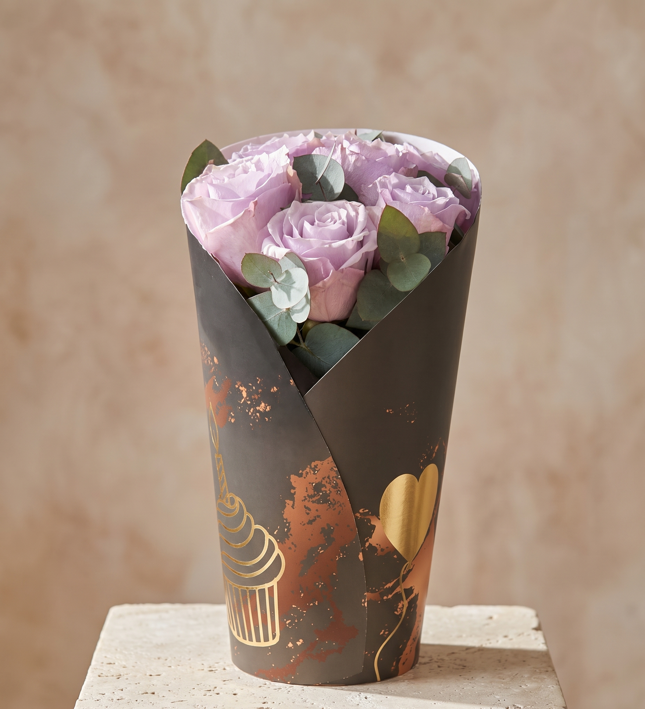 Joyeux Anniversaire, 7 Roses Lilas en Emballage Papier, Sept roses lilas et de l'eucalyptus, enveloppés à la main dans un cornet en papier à thème anniversaire, orné de motifs dorés de cupcake et de ballon.