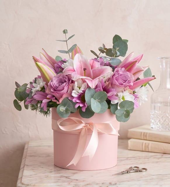 Rosa Lilien, Lila Rosen und Weiße Margeriten im Rosa Topf, Importierte rosa Lilien, zwei lila Rosen, weiße Margeriten und Ballerina-Kirschblüten mit Eukalyptus in einem rosa Topf mit Schleife, 25 bis 30 cm hoch.